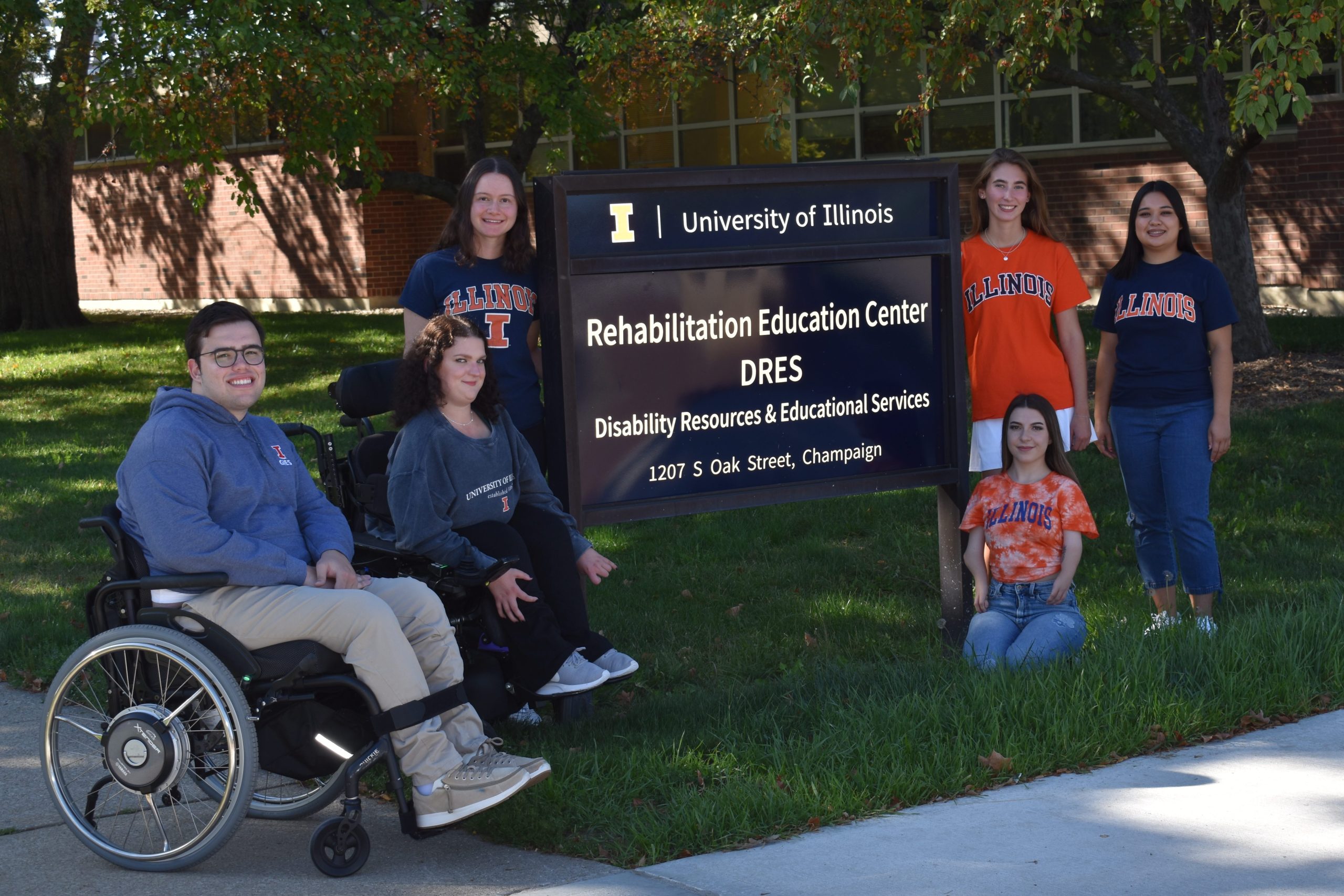6 people gather around sign outside of a building. Text on sign:
 University of Illinois
Rehabilitation Education Center
DRES
1207 S Oak Street, Champaign